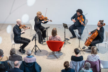 Vier Musikerinnen und Musiker mit Streichinstrumenten sitzen im Kreis und spielen, in ihrer Mitte eine Person in einem roten Sessel, am Rand sitzt Publikum.