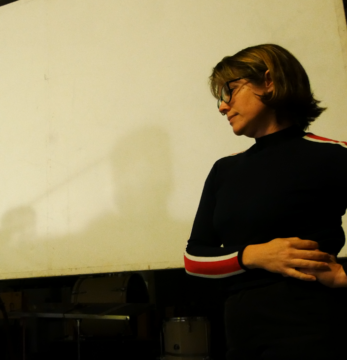 Eine Frau mit Brille und kinnlangen Haaren steht vor einer gelblichen Leinwand und schaut diese über die Schulter hinweg an. Es ist Heather Frasch.