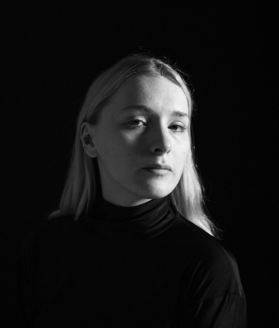 Schwarz-weiß-Portrait einer jungen Frau mit hellen, schulterlangen Haaren im schwarzen Rollkragenpullover, es ist Komponistin Kate Milligan.