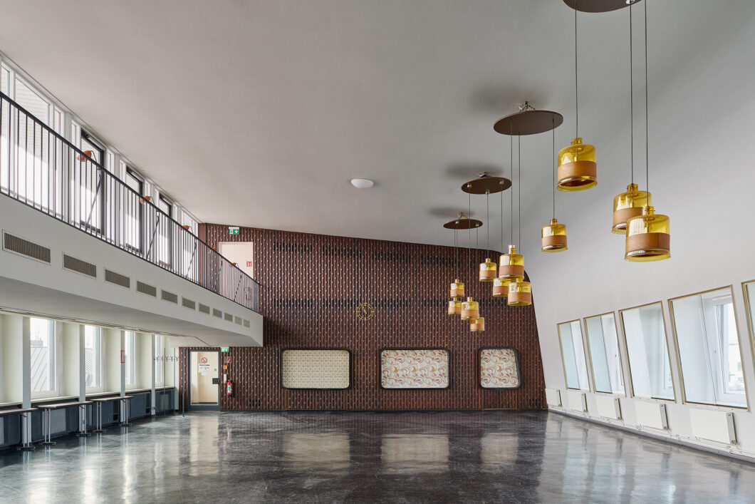 Ein leerer, lichtdurchfluteter Raum, rechts und links weiße Wände und Fenster, geradeaus eine braune Wand, ein Arrangement aus mehreren goldenen Hängelampen, es ist das Casino im InterCity Hotel