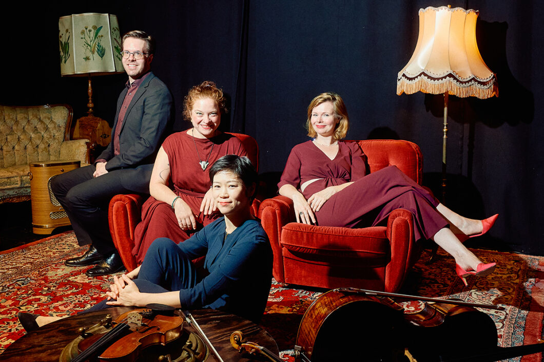 Ein Mann mit Brille und drei Frauen sitzen in einem Wohnzimmer, das im Retro-Look eingerichtet ist. Es ist das Flex Ensemble.