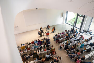 Von oben fotografiert: Menschen sitzen in einem hellen Raum in Reihen, eine Person sitzt vorn und vier Musiker*innen drumherum. Es ist ein Konzert des Treppenhausquartetts.