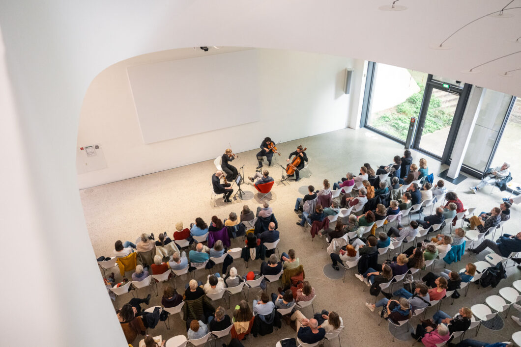 Von oben fotografiert: Menschen sitzen in einem hellen Raum in Reihen, eine Person sitzt vorn und vier Musiker*innen drumherum. Es ist ein Konzert des Treppenhausquartetts.