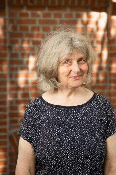Eine ältere Frau mit halblangen grauen Haaren und schwarzem, weiß gepunktetem T-Shirt steht vor einer roten Ziegelsteinwand und lächelt in die Kamera; es ist Astrid Schmeling.