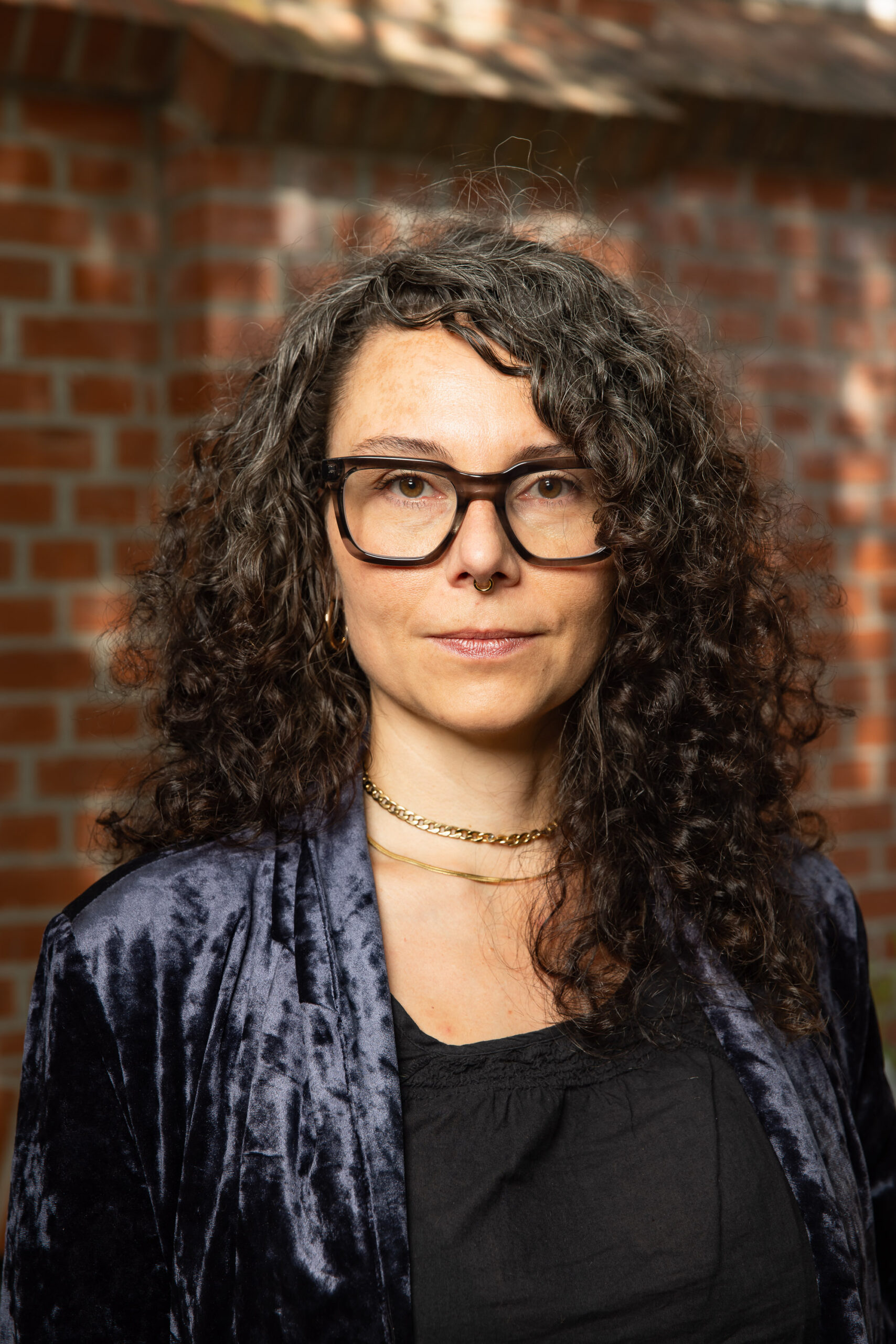 Eine Frau mit dunkelbraunen schulterlangen Locken, großer schwarzer Brille, blauem Samtjackett und schwarzem Shirt schaut in die Kamera, im Hintergrund eine rote Ziegelsteinwand; es ist Jacqueline Moschkau
