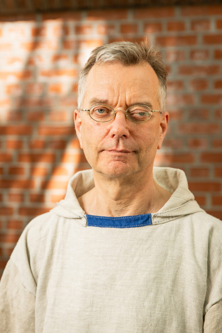 Ein Mann mit kurzen grauen Haaren und Brille in beigefarbenem Pullover steht vor einer roten Ziegelsteinwand: Komponist Joachim Heintz