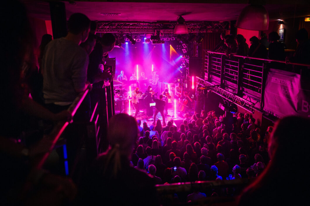 Eine pink erleuchtete Bühne mit einem klassischen Orchester, im Vordergrund Publikum. Es ist das Treppenhausorchester.