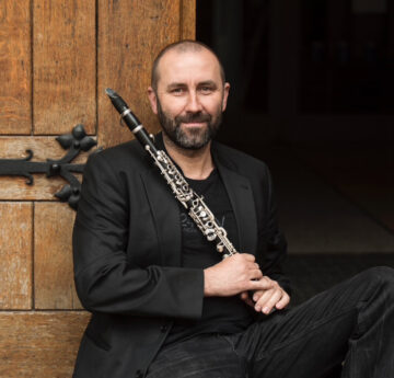 Ein Mann in schwarz mit einer Klarinette in der Hand sitzt an eine Holztür gelehnt. Es ist der Musiker Anton Säckl.