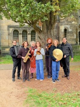 Menschen stehen in bunter Kleidung mit verschiedenen Instrumenten unter einem Baum