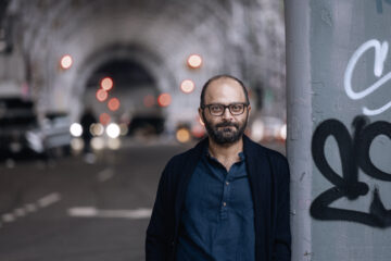 Ein Mann mit kurzen dunklen Haaren und Bart, trägt ein dunkelblaues Hemd und eine schwarze Strickjacke, steht unter einer Bogenbrücke. Es ist Komponist Ashkan Behzadi
