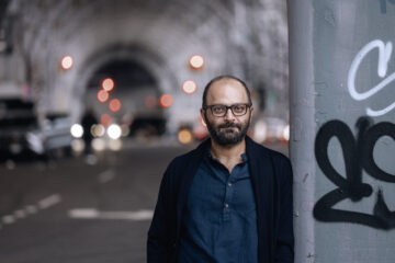 Ein Mann mit kurzen dunklen Haaren und Bart, trägt ein dunkelblaues Hemd und eine schwarze Strickjacke, steht unter einer Bogenbrücke. Es ist Komponist Ashkan Behzadi