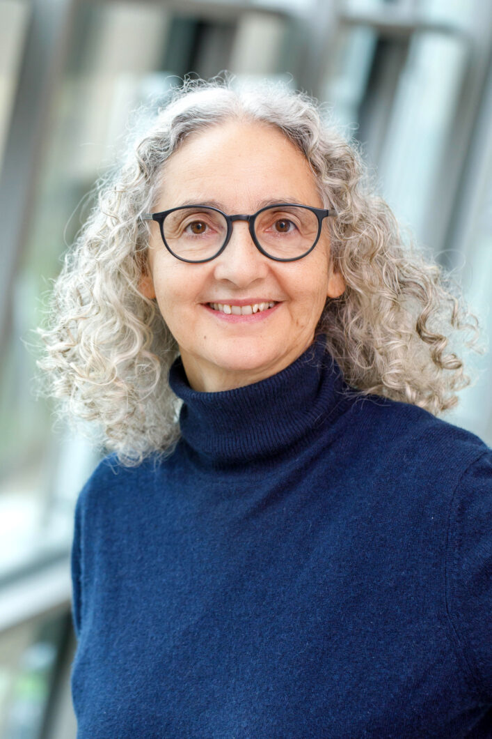 Eine Frau mit schulterlangen grauen Locken, dunkler runder Brille und blauem Rollkragenpullover lächelt in die Kamera, es ist Komponistin Caroline Bauckholt.