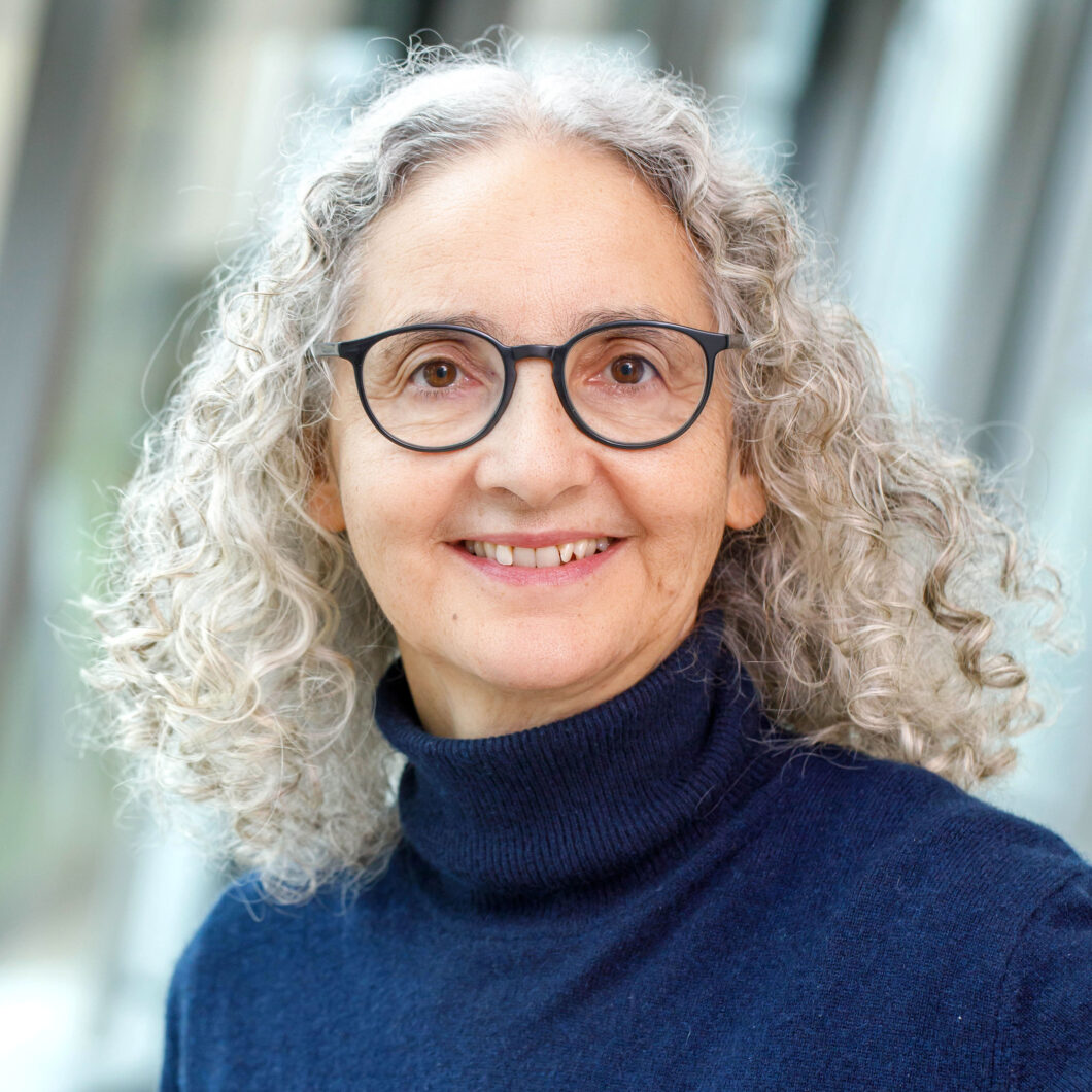 Eine Frau mit schulterlangen grauen Locken, dunkler runder Brille und blauem Rollkragenpullover lächelt in die Kamera, es ist Komponistin Carola Bauckholt.