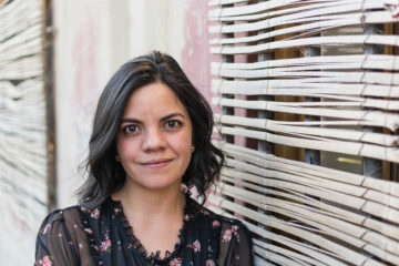 Eine junge Frau mit schwarzen halblangen Haaren lehnt an einer Wand, es ist Carolina Noguera