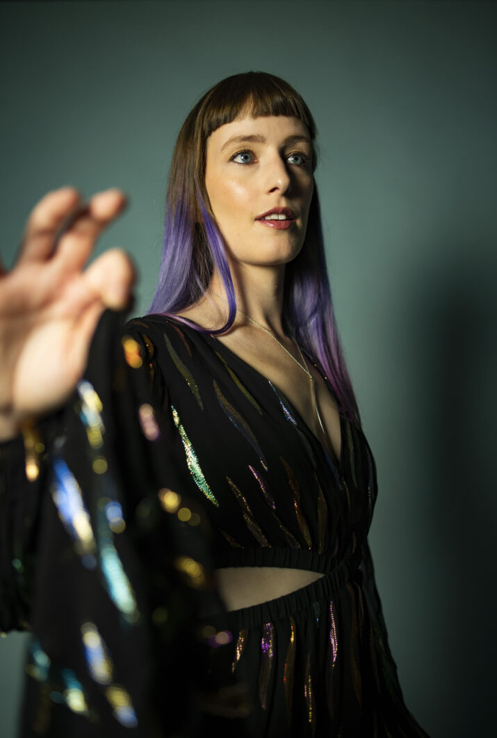 Eine junge Frau greift in Richtung Kamera, sie trägt ein schwarzes Kleid mit bunten Akzenten und braune lange Haare mit fliederfarbenen Spitzen. Es ist Sopranistin Charlotte Mundy.