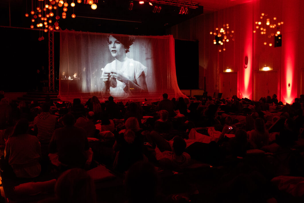 Ein rot erleuchteter Raum, auf der Leinwand die Projektion einer Frau in einem Schwarz-Weiß-Film.