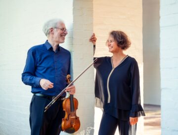 Ein Mann, graue kurze Haare und dunkelblaues Hemd, mit Geige in der Hand steht neben einer Frau in schwarzer Bluse, beide lächeln sich an, es ist das Duo Expressionen.