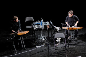 Auf einer Bühne stehen Stühle, Laptops, Perkussionsinstrumente, links und rechts sind zwei Musiker in Aktion.