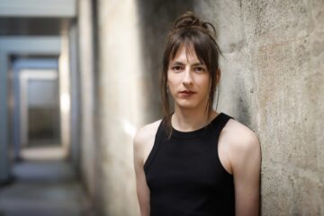 Porträtfoto einer Person mit dunklen, hochgesteckten Haaren, mit ärmellosem Top, angelehnt an eine Wand. Es ist Komponist:in Eloain Lovis Hübner (dey/sie).