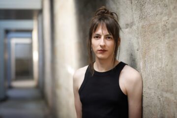 Porträtfoto einer Person mit dunklen, hochgesteckten Haaren, mit ärmellosem Top, angelehnt an eine Wand. Es ist Komponist:in Eloain Lovis Hübner (dey/sie).