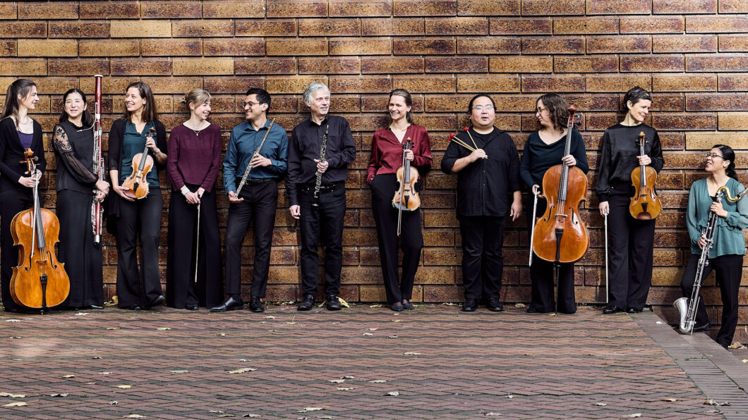 Elf Musikerinnen und Musiker stehen mit verschiedenen klassischen Instrumenten in der Hand an eine Backsteinwand gelehnt und unterhalten sich. Es ist das Ensemble Horizonte