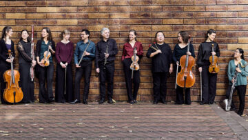Elf Musikerinnen und Musiker stehen mit verschiedenen klassischen Instrumenten in der Hand an eine Backsteinwand gelehnt und unterhalten sich. Es ist das Ensemble Horizonte