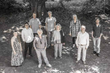 Eine Gruppe Menschen steht, gekleidet in Grau und Weiß, unter Bäumen, durch die Licht fällt.