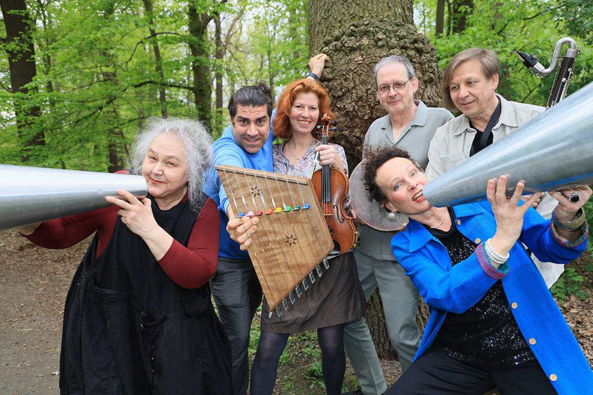 Eine Gruppe aus sechs Menschen steht in einem Wald und hält verschiedene Instrumente in die Kamera und lacht. Es ist Ensemble Megaphon.