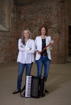 Zwei Frauen in blauen Shirts mit schwarzen Blaisern stehen an einer Treppe vor einer Backsteinwand, die linke hat ein Akkordeon neben sich, die rechte hält eine Schalmei in den Händen. Es ist das Ensemble Mixtura.