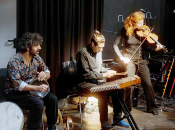 Drei Musiker*innen mit (von links nach rechts) einer Trommel, einer Santur und einer Geige spielen auf einer Bühne.