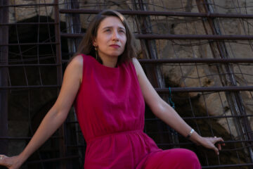 Eine junge Frau mit halblangen braunen Haaren sitzt zurückgelehnt in einem weinroten Kleid vor einer Backsteinwand. Es ist Pianistin Fidan Aghayeva-Edler.