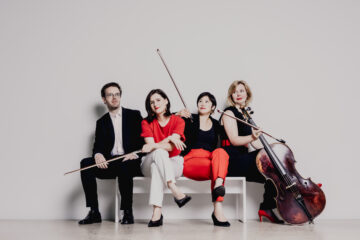 Vier Musikerinnen und Musiker sitzen mit ihren Instrumenten auf einer Bank vor einer weißen Wand.