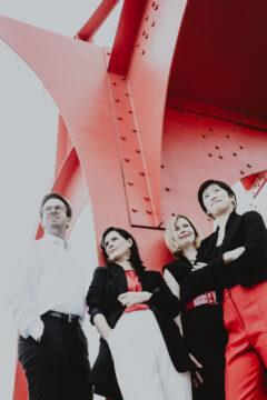 Schwarz-weiß Foto, teilweise rot eingefärbt: Ein Mann und drei Frauen stehen mit verschränkten Armen vor einem Metallkonstrukt. Es sind die Musiker*innen des Flex Ensembles.