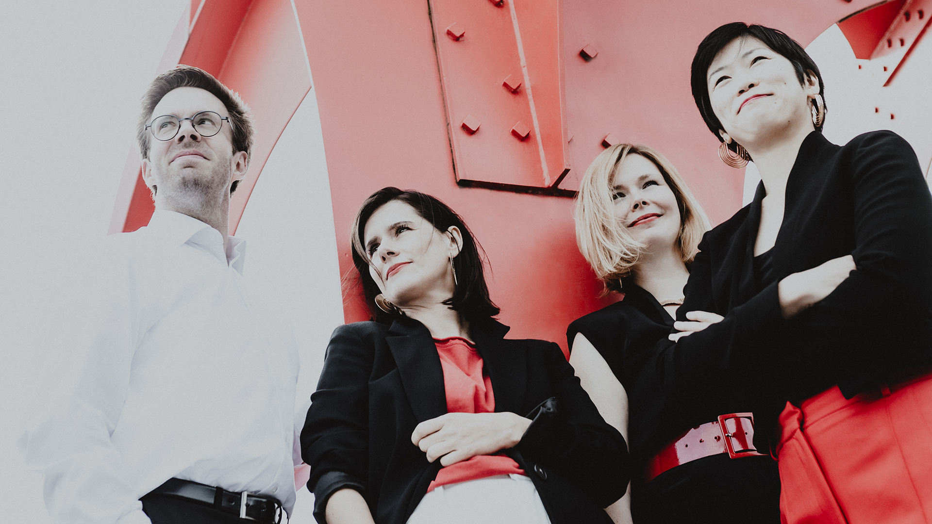 Schwarz-weiß Foto, teilweise rot eingefärbt: Ein Mann und drei Frauen stehen mit verschränkten Armen vor einem Metallkonstrukt. Es sind die Musiker*innen des Flex Ensembles.