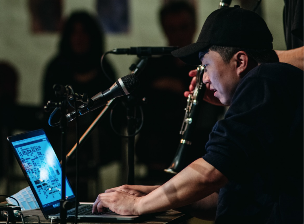 Ein Mann mit schwarzem Basecap gibt etwas in einen Laptop ein, vor ihm ein Mikrofon, im Hintergrund Publikum. Es ist Komponist der koreanische Hunjoo Jung.
