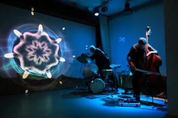Zwei Personen auf einer Bühne neben einer Videoprojektion, die aussieht wie eine Blume, sie spielen Schlagzeug und Kontrabass, der Raum ist in blaues Licht getaucht.