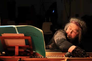 Eine grauhaarige Frau sitzt an einem Flügel und greift beim Spielen hinein, im Vordergrund das Notenheft.