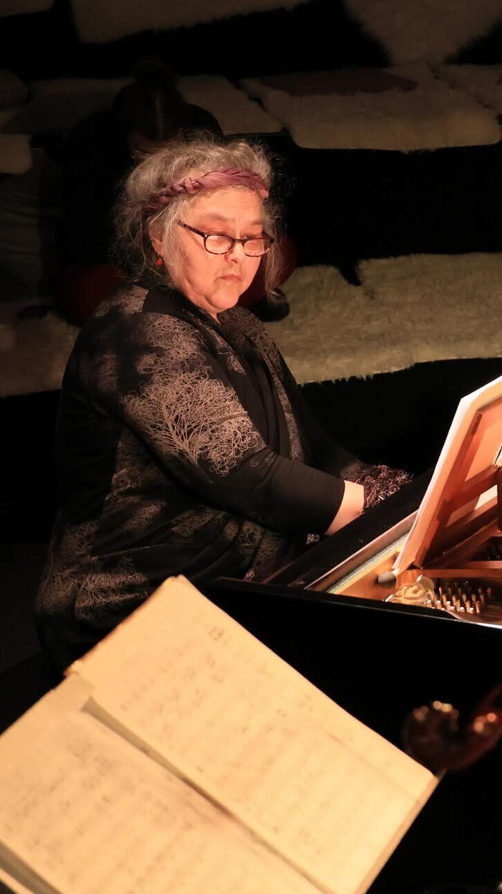 Eine grauhaarige Frau mit Brille sitzt an einem Flügel und spielt, im Vordergrund ein Notenheft auf einem Notenständer. Es ist die Musikerin Tatjana Prelević.