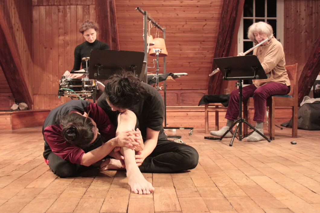 Zwei Menschen hocken ineinander verschlungen auf einem Holzfußboden, im Hintergrund eine Frau, die Querflöte spielt, und eine Frau, die Percussioninstrumente spielt.