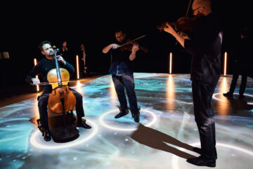 Ein Cellist und zwei Geiger spielen in einem dunklen Raum, der Fußboden leuchtet türkis und weiß.
