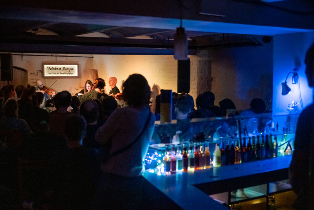 Eine Theke, bunt beleuchtete Getränkeflaschen, die Schattenrisse von Menschen, im Hintergrund Musiker*innen hell beleuchter – es ist der hannoversche Kellerclub Feinkost Lampe