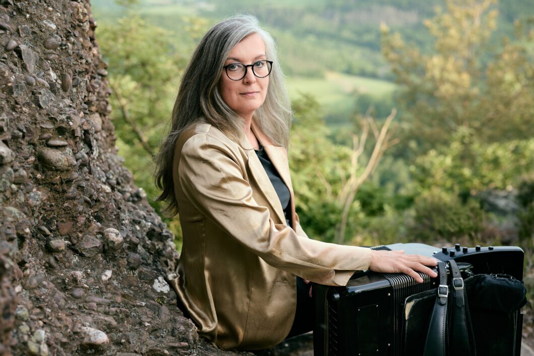 Eine Frau mit langen grauen Haaren, dunkler runter Brille und goldenem Jackett sitzt mit einem Akkordeon neben sich an einem Baum. Es ist die Musikerin Margit Kern.