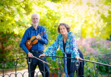 Ein Mann im blauen Hemd mit Geige und eine Frau in blauer Bluse stehen an ein Geländer gelehnt, im Hintergrund ein grünender Garten.