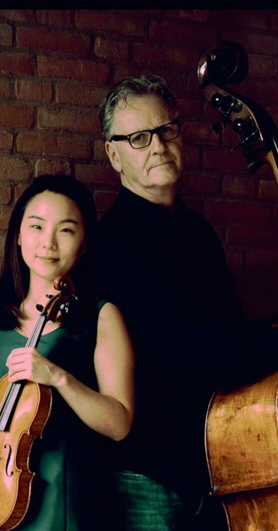 Eine junge Frau mit Geige in der and lehnt seitlich an einem älteren Mann mit Kontrabass. Es sind die Musiker*innen Seayoung Kim und olger Michalski.