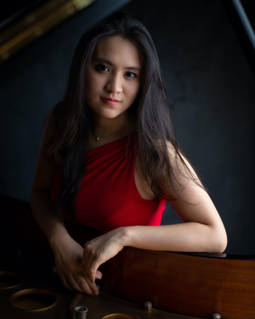 Eine junge Frau mit langen dunklen Haaren im roten Oberteil lächelt in die Kamera. Es ist Pianistin Shan-Chi Hsu