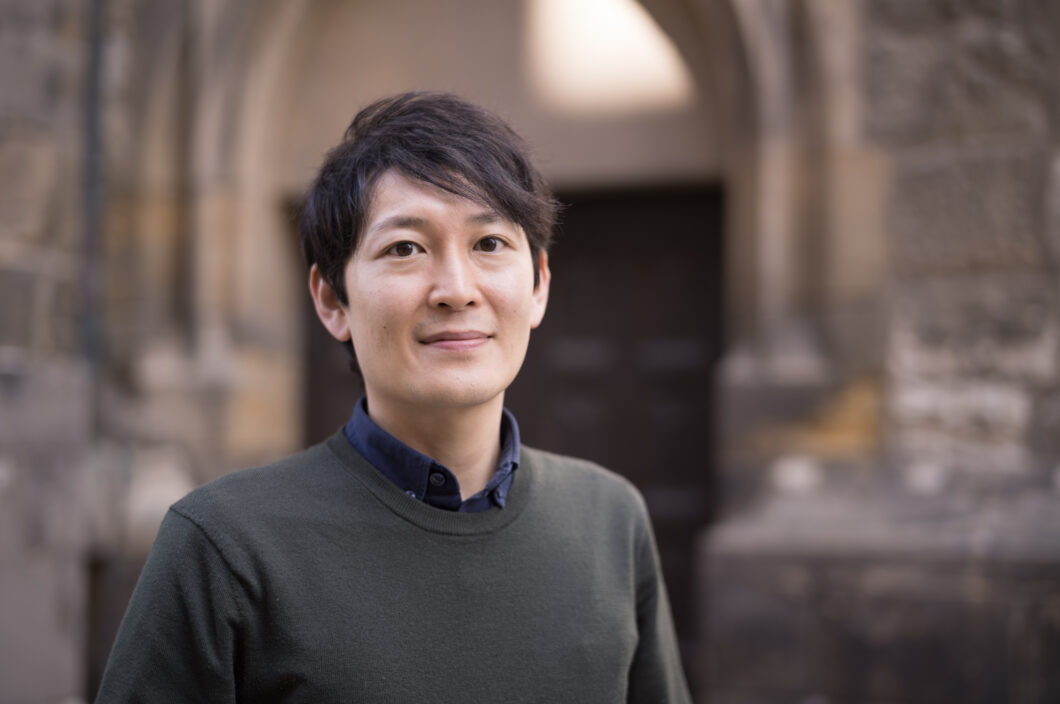 Ein Mann mit schwarzen kurzen Haaren lächelt in die Kamera. Es ist Komponist Tomoya Yokokawa.