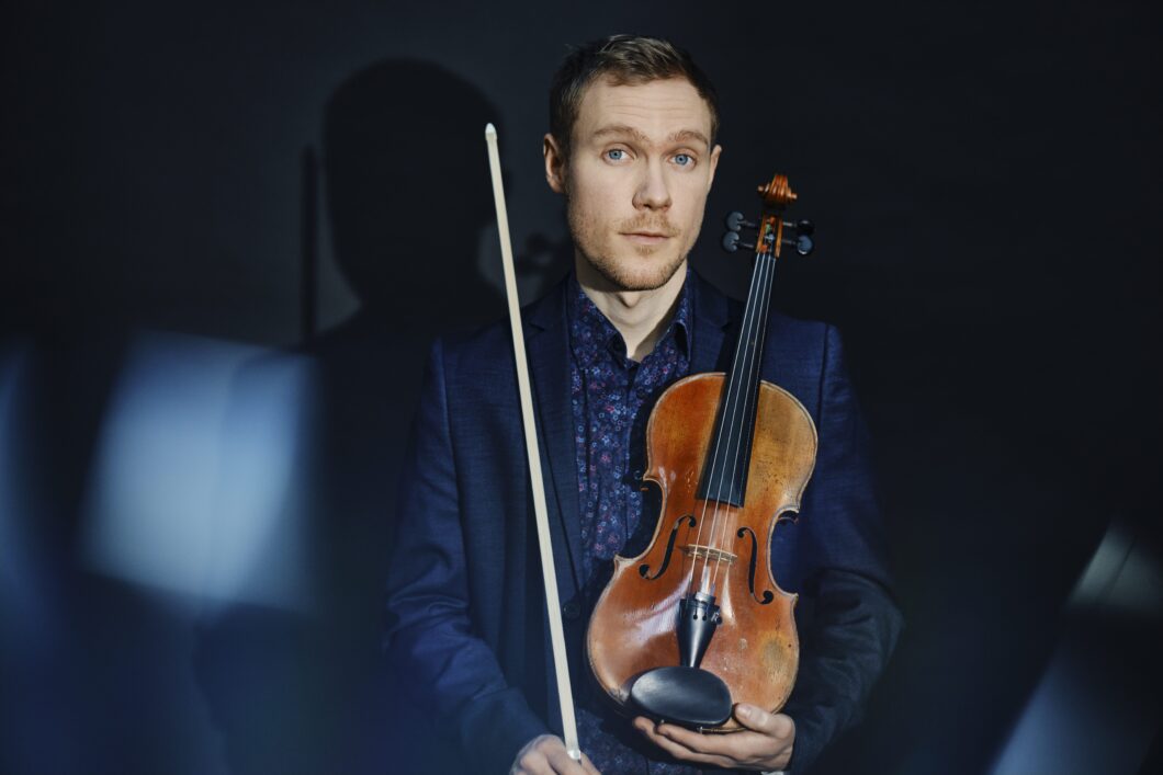 Ein junger Mann mit kurzen rotbraunen Haaren, blaues Hemd und blaues Jackett, hält eine Violine vor der Brust. Es ist Geiger Adam Woodward.