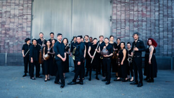 Eine Gruppe Menschen, schwarz gekleidet mit Instrumenten, steht vor einem Metalltor; es ist das Ensemble Asambura.