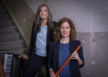 Zwei Frauen in blauen Shirts mit schwarzen Blaisern stehen an einer Treppe vor einer grauen Wand, die linke hat ein Akkordeon neben sich, die rechte hält eine Schalmei in den Händen. Es ist das Ensemble Mixtura.