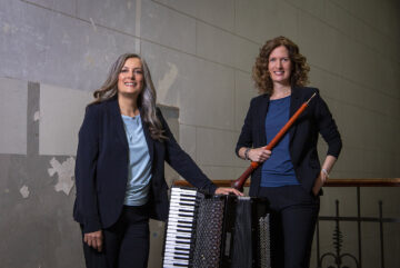 Zwei Frauen in blauen Shirts mit schwarzen Blaisern stehen vor einer grauen Wand, die linke hat ein Akkordeon neben sich, die rechte hält eine Schalmei in den Händen. Es ist das Ensemble Mixtura.